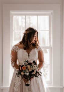 One Fine Day Bride, bride in a long sleeve wedding dress holding her bouquet in front of a window