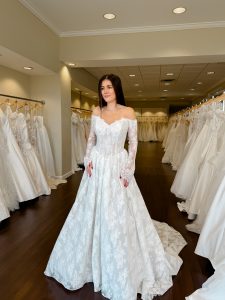 A bride with long dark hair wears a long sleeve off the shoulder wedding dress in an all over chantilly lace standing between rows of bridal gowns at One Fine Day Bridal in Fort Wayne, Indiana.