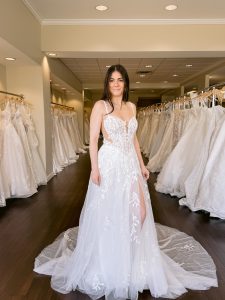 A petite bride with long dark hair wears lace and tulle wedding dress with thin lace straps and side cut outs and a leg slit standing between rows of bridal gowns at One Fine Day Bridal in Fort Wayne, Indiana.