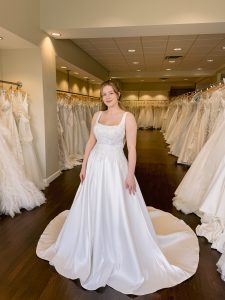 A bride with blonde hair wears a square neck wedding dress in ivory with lace and beading on the bodice and side cut outs standing between rows of bridal gowns at One Fine Day Bridal in Fort Wayne, Indiana.