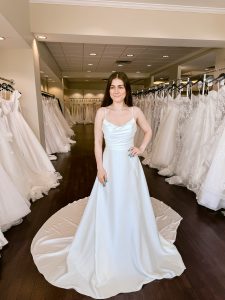 A petite bride with dark hair wears a satin aline wedding dress with spaghetti straps and pearl beading along the neckline standing between rows of bridal gowns at One Fine Day Bridal in Fort Wayne, Indiana.