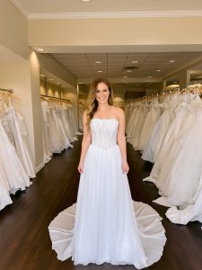 A petite bride wears a strapless wedding dress with a lace bodice and crinkle chiffon skirt in ivory standing between rows of bridal gowns at One Fine Day Bridal in Fort Wayne, Indiana.