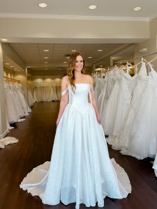 A petite bride wears a strapless jacquard ballgown wedding dress with narrow straps that swoop in ivory standing between rows of bridal gowns at One Fine Day Bridal in Fort Wayne, Indiana.