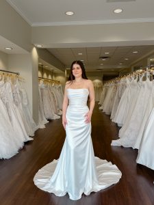 A petite bride with long dark hair wears a fitted strapless silk satin wedding dress in ivory with ruching and draping throughout the bodice and skirt standing between rows of bridal gowns at One Fine Day Bridal in Fort Wayne, Indiana.