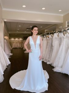 A petite bride with a ponytail wears a crepe fitted wedding dress with vneckline and scattered pearls and beads on neckline in ivory standing between rows of bridal gowns at One Fine Day Bridal in Fort Wayne, Indiana.