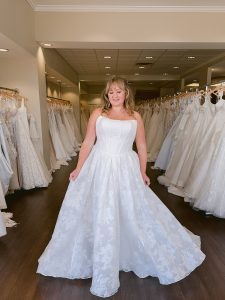 A mid size bride wears a strapless jacquard wedding dress in ivory standing between rows of bridal gowns at One Fine Day Bridal in Fort Wayne, Indiana.
