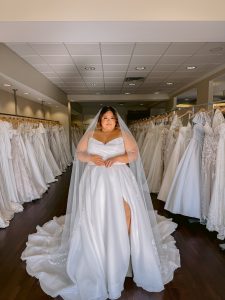 A plus size bride with wears a strapless organza wedding dress in ivory with a leg slit and cathedral length veil standing between rows of bridal gowns at One Fine Day Bridal in Fort Wayne, Indiana.