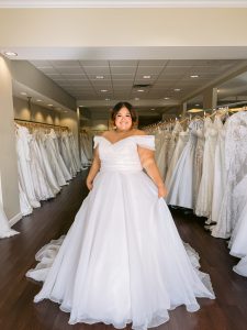 A plus size bride wears an off the shoulder organza ballgown wedding dress in blush standing between rows of bridal gowns at One Fine Day Bridal in Fort Wayne, Indiana.